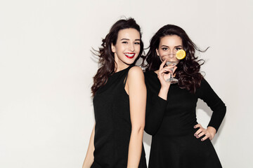  Two brunette sisters with long hair drink an alcoholic cocktail and wine. Festive girls in evening dress laugh and smile. A bottle of wine. White background.