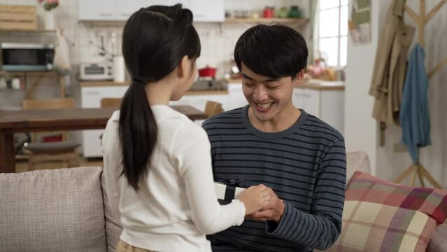 view from behind Asian little girl walking up to her dad and showing him a surprise gift in living room at home. happy father embracing her daughter for the sweet act on father's day