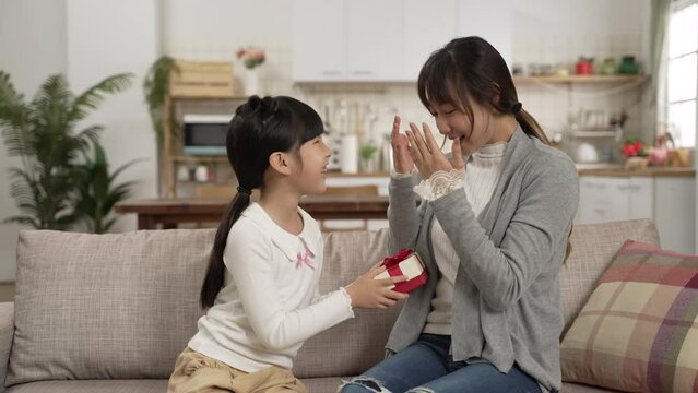 slow motion of surprised asian mother embracing daughter for getting mother&rsquo;s day gift at home. the girl showing present from behind as her mom is covering eyes
