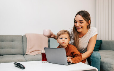 Fototapeta premium Happy middle age woman playing with her little son at home. They are using smart phone and laptop computer for playing, learning and online surfing.