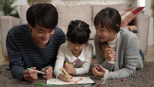 slow motion of sweet asian family of three dad, mom and schoolgirl daughter drawing on paper with color pens on living room floor at home