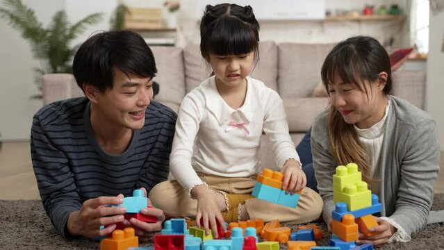 Asian father and mother lying on floor playing colorful building blocks with their schoolgirl daughter at home. they race with each other stacking up the pieces