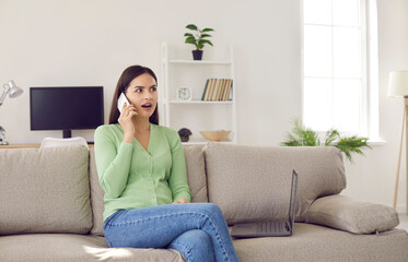 Young girl sharing news and learning latest gossip on phone. Beautiful Caucasian woman sitting on sofa at home and talking to her friend on cellular phone with surprised, shocked face expression