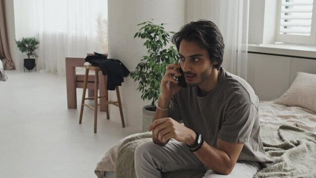 Medium long of young Middle Eastern man in casual clothes sitting on bed in modern apartment, talking on cellphone in morning