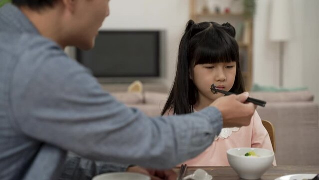 Selective Focus Of Unhappy Asian Daughter Turning Head While Refusing Food His Father Is Feeding Her During Lunch Time In Dining Room At Home