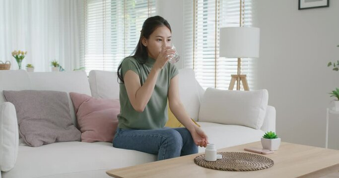 Asia people young woman happy relax smile sit at home sofa drink water take birth control pill pouring folic preconception drug bottle or weight loss diet capsule. Body skin health care sick relief.