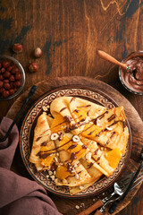 Crepes suzette with oranges and chocolate. Thin crepes with chocolate spread, hazelnuts and orange slices fruit in plate for breakfast on old dark wooden rustic table background. Copy space. Top view