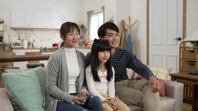 Beautiful smiling Asian family of three sitting together watching tv at home. Mom tuning channels with controller and dad explaining to their daughter with finger pointing
