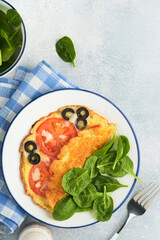 Omelette with tomatoes, black olives, cottage cheese and green spinach herbs on white plate on grey concrete background. Delicious breakfast. Healthy breakfast food. Top view.