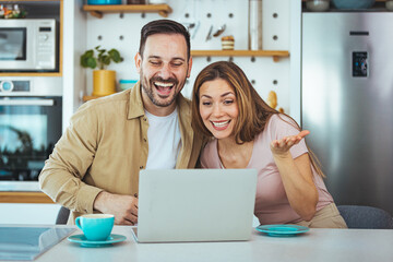 Happy young family couple bonding laughing using laptop looking at screen sit at table, smiling husband and wife buying choosing goods online doing internet shopping order with delivery at home