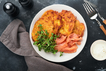 Potato pancakes. Fried homemade potato pancakes or latkes with cream, green onions, microgreens, red salmon and sauce in rustic plate on old black table background. Rustic style. Top view.