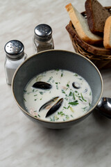 A small portion of milk clam chowder with mussels and dill, bread basket aside, light marble table