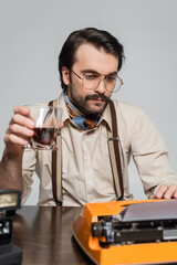 journalist in eyeglasses looking at paper in typewriter while holding glass of whiskey isolated on grey.