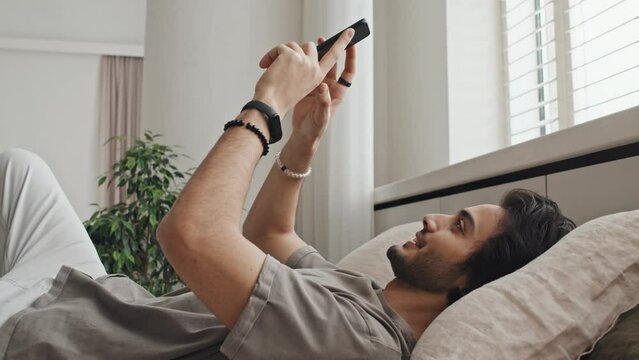 Medium long side-view of young cheerful Middle Eastern man in casual clothes lying on bed, using smartphone in morning, scrolling social media feed, smiling