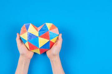 Child hands holding colorful polygonal paper origami heart on light blue paper background. World...