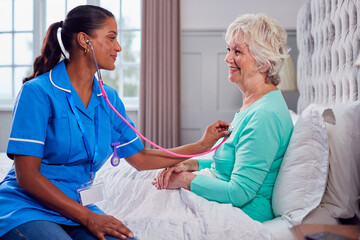 Female Care Worker In Uniform Listening To Chest Of Senior Woman At Home In Bed With Stethoscope