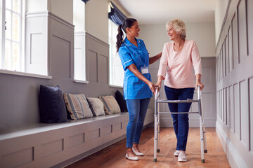 Senior Woman At Home Using Walking Frame Being Helped By Female Care Worker In Uniform