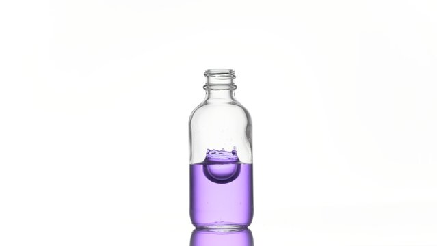 Drop of niacinamide falls down into medical bottle with purple transparent liquid on white background and makes crawn | Abstract skincare cosmetics with niacinamide formulation concept