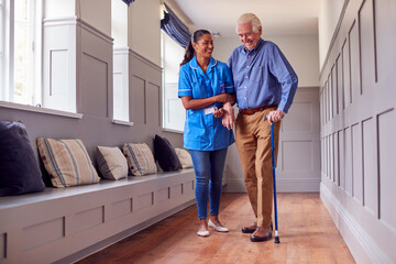 Senior Man At Home Using Walking Stick Being Helped By Female Care Worker In Uniform