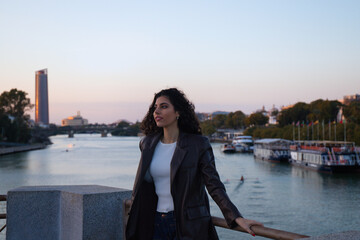 Young and beautiful woman with dark and curly hair is sightseeing in Seville. In the background the river guadalquivir and part of the city in the golden hour. Tourism and holidays concept.