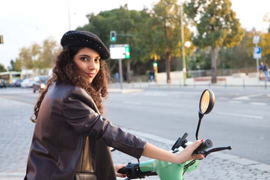 Young and beautiful woman with dark and curly hair is sightseeing in seville while driving an electric motorbike to move around the city. Renewable and non-polluting energy. Tourism and holidays