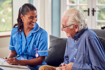 Senior Man At Home Talking To Female Nurse Or Care Worker In Uniform Making Notes In Folder