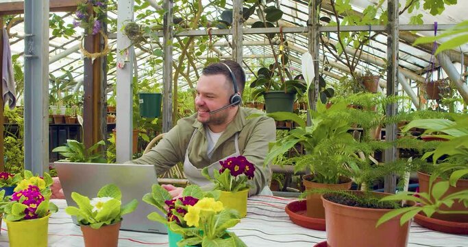 A young greenhouse worker during an online video call - he sitting in front of laptop and telling customers about the grown flowers to the camera and actively gesticulates.