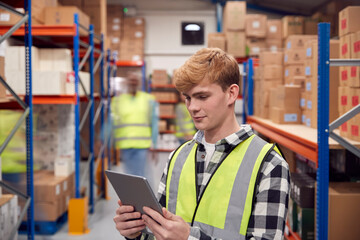 Male Intern With Digital Tablet In Busy Modern Warehouse With Staff In Background
