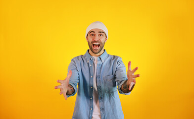 Super surprised. Studio shot of smiliing handsome man in jeans and white hat on yellow background. Tatoo and beard.