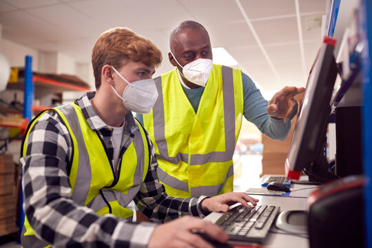 Male Intern With Supervisor Wearing Face Masks Working In Busy Modern Warehouse On Computer Terminal