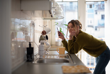 Woman standing in the kitchen and drinking cup of tea while using smartphone