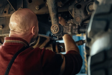 Bald car mechanic or mechanic sits under machinery and repairs engine suspension of car in...