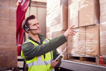 Male Worker Wearing Headset At Freight Haulage Business With Truck Being Loaded By Fork Lift 