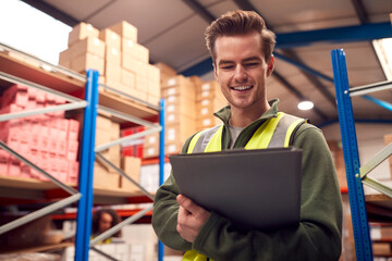 Male Worker Wearing High-Vis Vest Inside Busy Warehouse Checking Stock On Shelves Using Clipboard