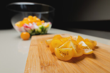 Sliced yellow cherry tomatoes on a wooden board in shallow depth of field against the background of a bowl with veggie salad ingredients