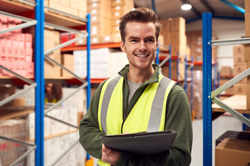 Male Worker Wearing High-Vis Vest Inside Busy Warehouse Checking Stock On Shelves Using Clipboard