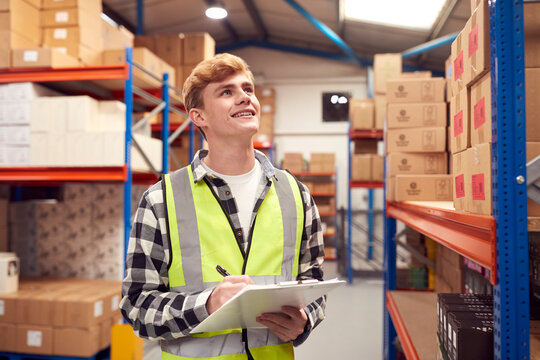 Male Intern Working Inside Warehouse Checking Stock On Shelves Using Clipboard