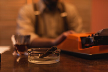 ashtray with cigar near typewriter machine and blurred man on background.