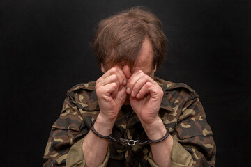 A military soldier or officer in handcuffs hides his face against a dark background. War criminal,...