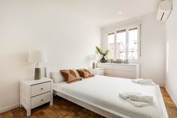 Fototapeta na wymiar Bedroom with white wooden bedside tables, long dresser with many matching drawers under the window and king size bed with cushions and oak floorboards
