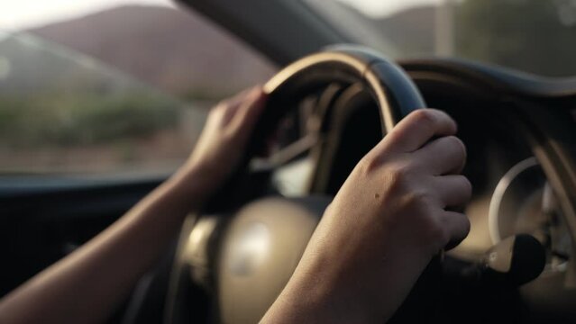 Female hands on steering wheel driving in a car during sunset or sunrise. Sun shining through vehicle windows as car going down the highway. Woman driving the car on the road