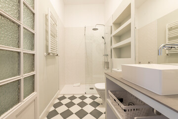 Bathroom with white and gray built-in furniture, porcelain sink, shower cabin with glass partition and checkered tiled floor