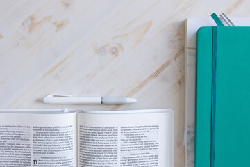 Border of open bible, teal journal and devotional and white pen on a white wood background with...