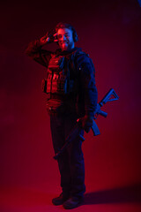 Fototapeta na wymiar a fighter in equipment, body armor, headphones holds a machine gun, aims at the target, hits the target through the sight. military business concept. red background. blue-red light