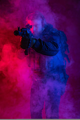 a fighter in equipment in smoke, body armor, headphones holds a machine gun, aims at the target, hits the target through the sight. military business concept. red background. blue-red ligh