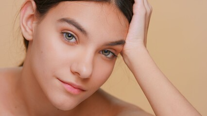 Close-up beauty portrait of young woman with pure skin who looks at camera props his head up with his hand on beige background