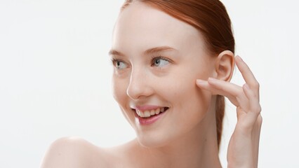 Obraz premium Slim redhead Caucasian woman touches her cheek, looks aside and smiles widely on white background | Moisturizer commercial