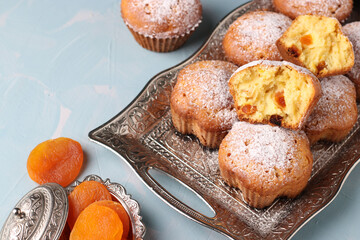 Homemade muffins with dried apricots sprinkled with icing sugar on a metal tray on a light blue background