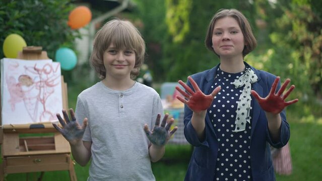 Caucasian boy and girl showing colorful palms looking at camera smiling standing outdoors. Portrait of joyful confident talented children posing in slow motion painting in park. Lifestyle and art