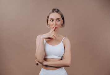 Thoughtful fit woman with beautiful face complexion holding finger on lips, looking away, thinking over solution. Model in white sport top and leggings posing on beige background.
